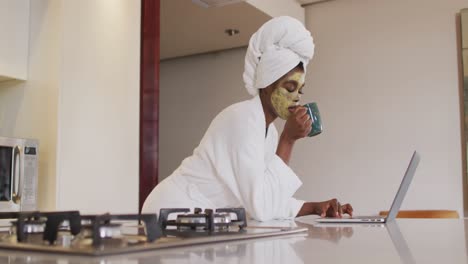 Mujer-Afroamericana-Atractiva-Con-Mascarilla-Aplicada-Tomando-Café-Y-Usando-Una-Computadora-Portátil-En-La-Cocina