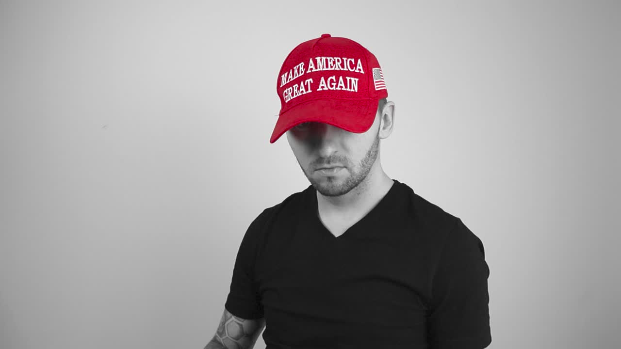 Grayscale footage of a tattooed man putting on a red MAGA Make America Great Again hat in slow motion and pointing towards the camera with his finger with confidence and approval. The man is happy.