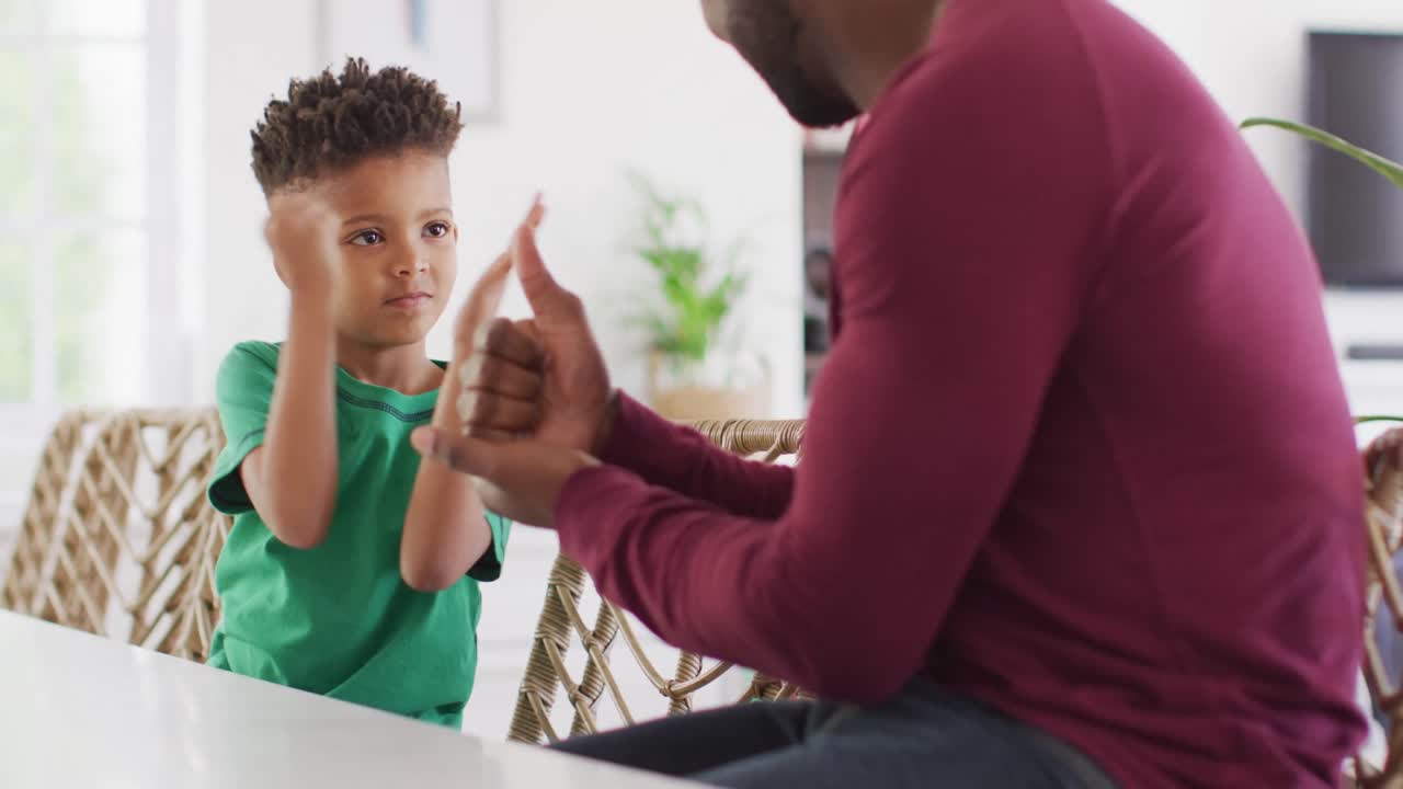 Happy african american man and his son sitting at table and speaking sign language