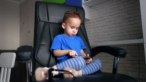 dark-haired male toddler in blue t-shirt sits with his legs folded in a big office chair. cute kid taps with his finger by the screen of smartphone. low angle view.
