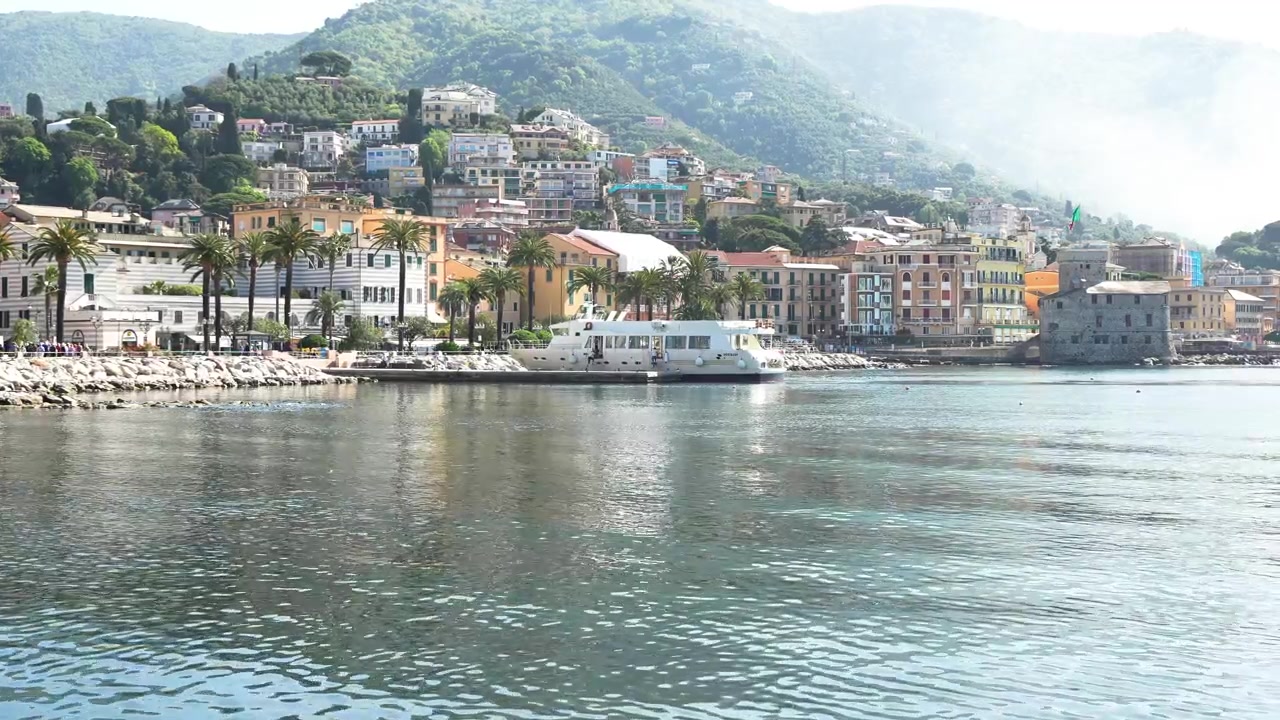 Inquadratura statica grandangolare del pittoresco lungomare di Rapallo, con colorati edifici liguri, una passeggiata fiancheggiata da palme, acque calme del porto e rigogliose colline verdi sotto la luce del giorno.