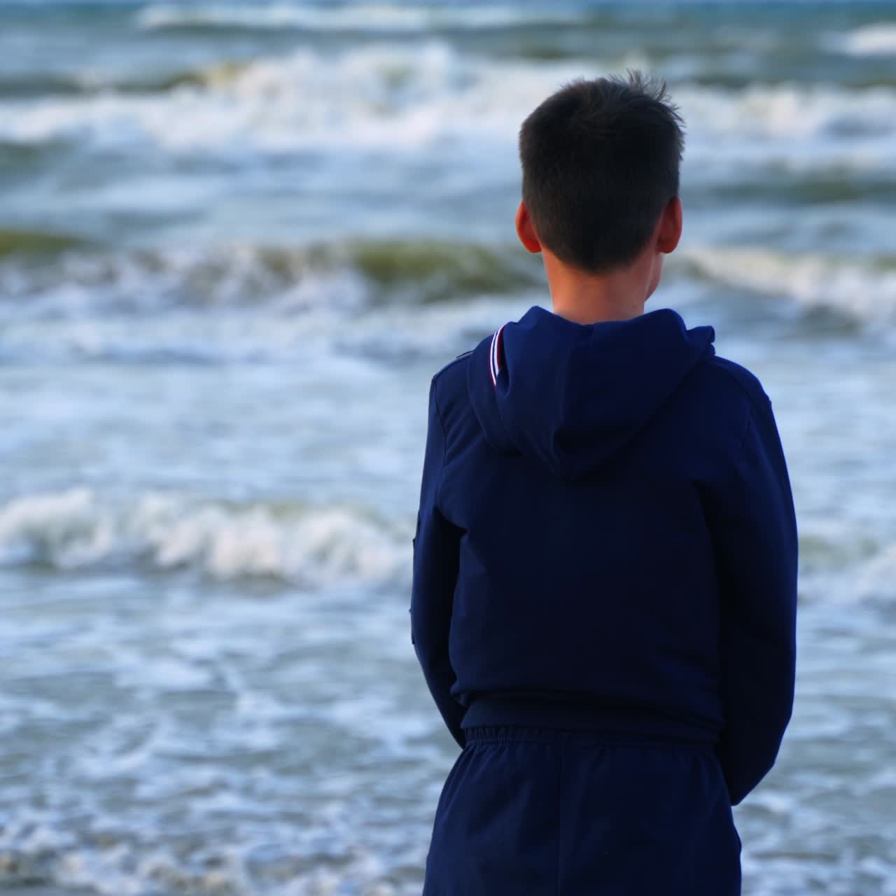 Back view of a boy near the blue sea. Teenage boy standing on the beach and looking on the sea water. Child dreaming on the ocean shore.