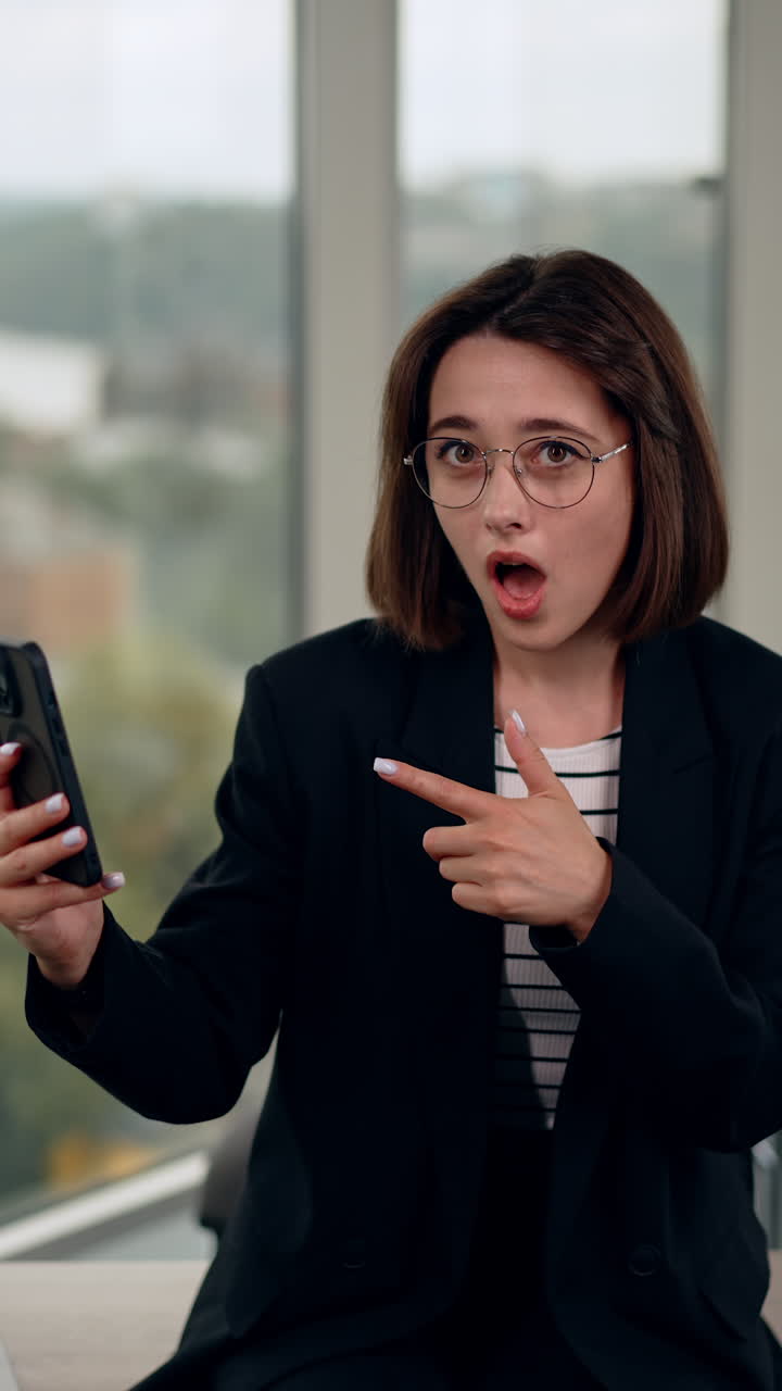 Attractive dark-haired lady looking at her phone with astonishment. Woman points at her gadget with mouth dropped. Vertical video.