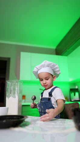 caucasian toddler kid smudged with flour stands at the table. low angle view at the kid holding a spoon and shaking his head with a chef cap. vertical video.