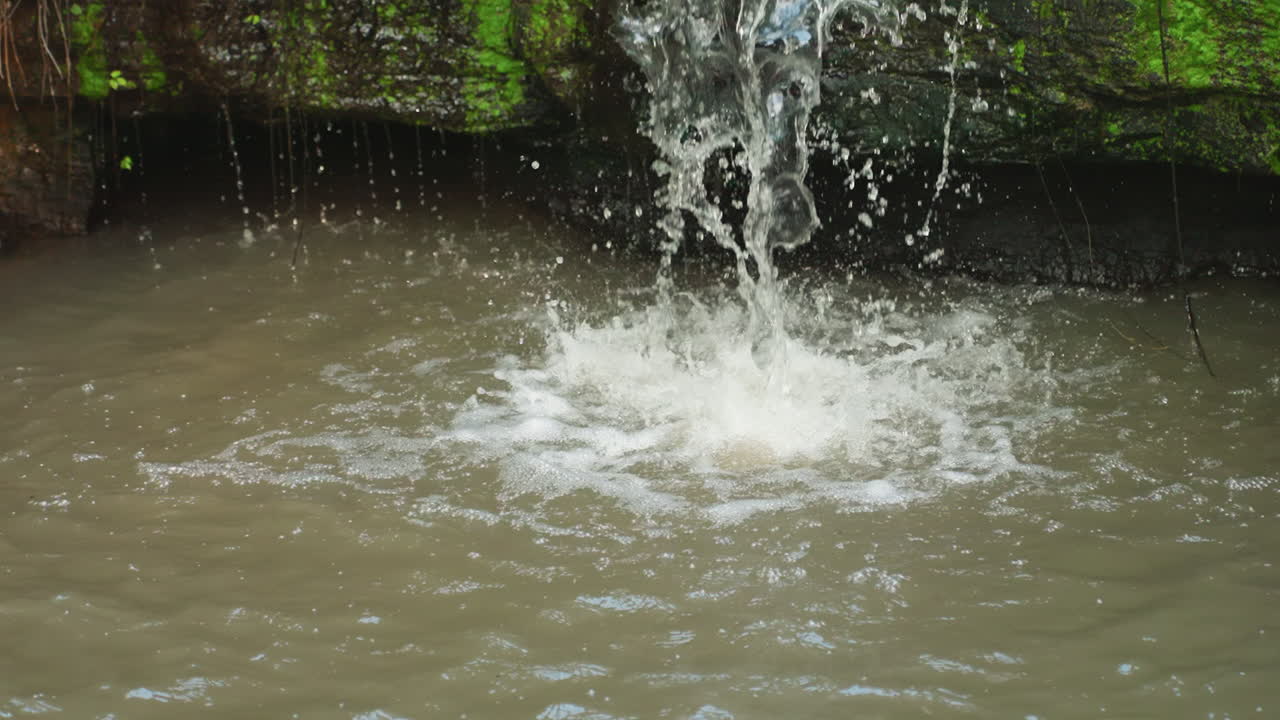 Air terjun jarak dekat ke kolam berlumpur, gua berlapis lumut dengan percikan bergejolak dan sedimen yang naik, tepian batu basah menunjukkan erosi, riak bertekstur dan suasana lembap di sekitarnya.