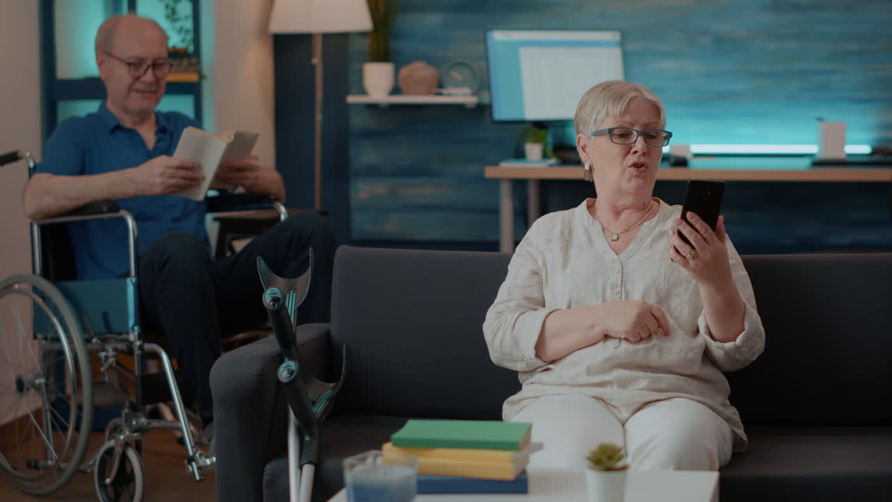 Senior Couple at Home: Woman Using Smartphone, Man Reading in Wheelchair