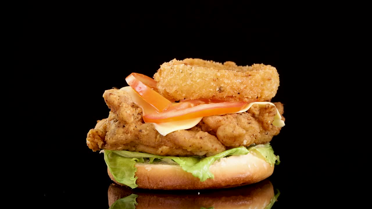 A crispy fried chicken burger with lettuce, tomato, cheese, and onion ring drops onto a reflective black surface in dramatic studio lighting