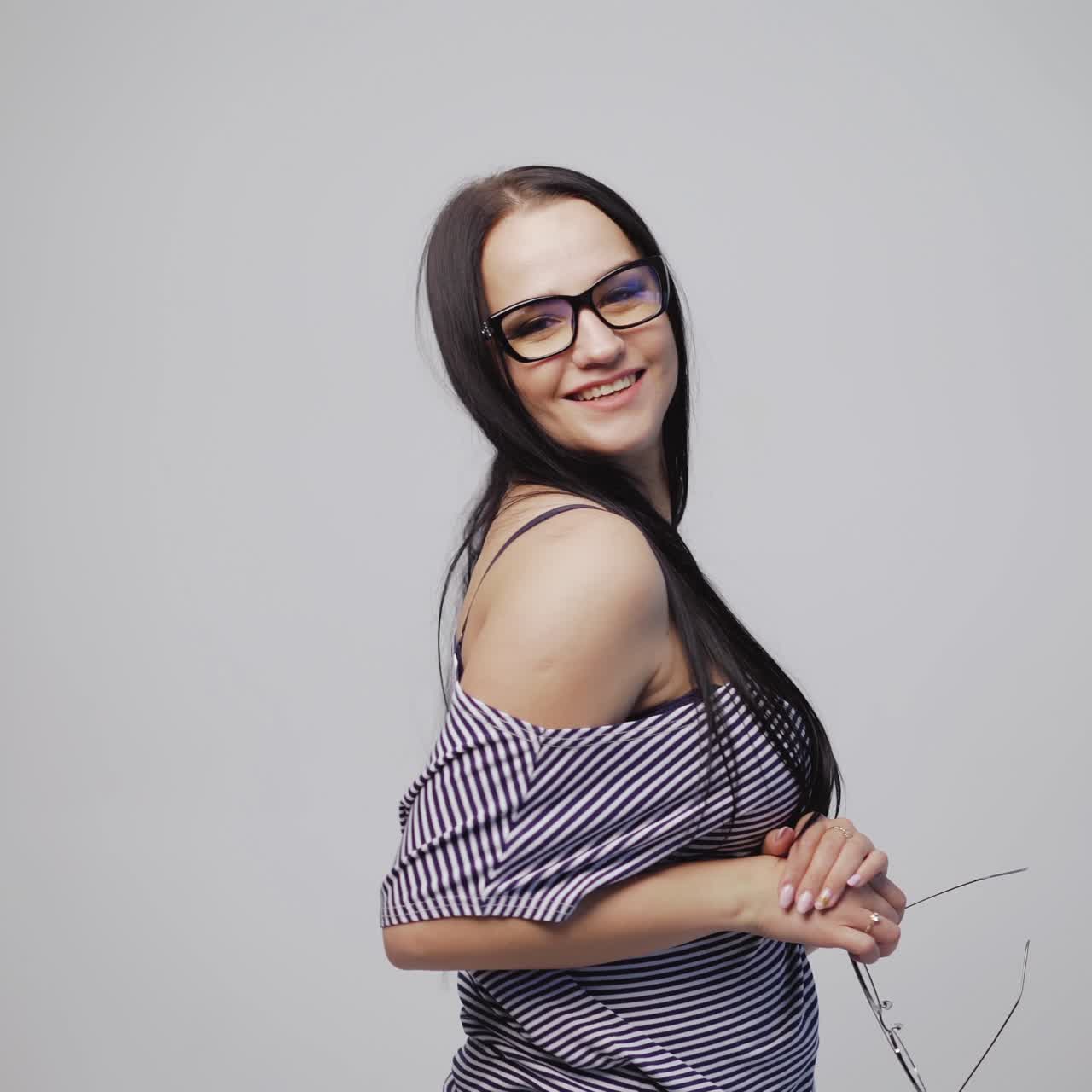 woman in a striped dress is choosing between two eyeglasses and putting on one of it in the studio
