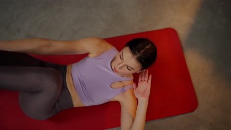 from above close up a confident brunette girl in a lilac top and on a red sports carpet goes in for sports and trains her abdominal muscles in a modern apartment