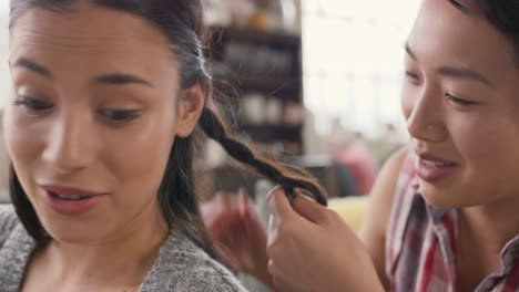 Multi-racial-girl-friends-braiding-hair-smiling-using-mobile-phone-hanging-out-on-sofa