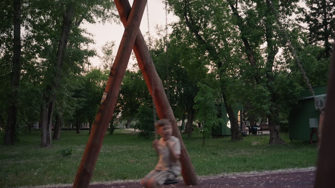 top down view of wooden swing structure and chains with children playing joyfully at dusk in lush tree lined park showcasing motion blur against serene evening sky with ambient light through leaves