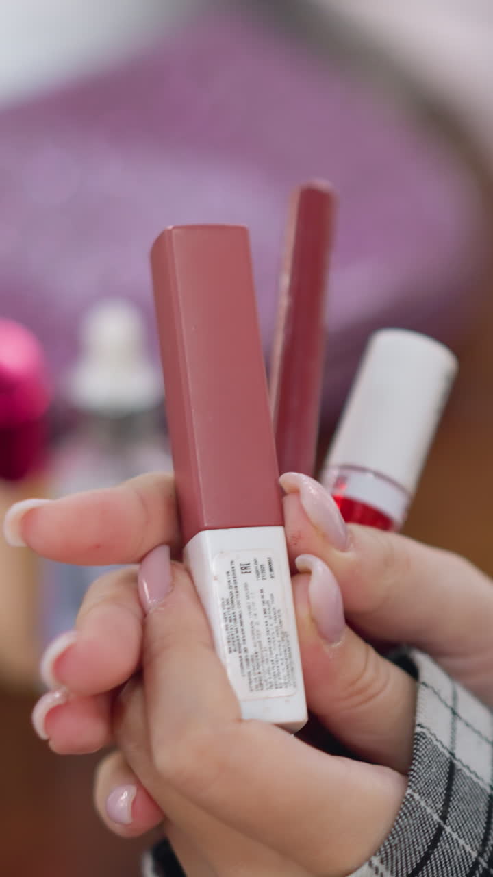 primer plano de unas manos sosteniendo un pintalabios malva y un brillo de labios rojo, uñas rosas con manicura, manga de cuadros, mano femenina inspeccionando un tubo de marca blanca, suave movimiento de muestra en la yema del dedo, iluminación interior cálida, íntimo