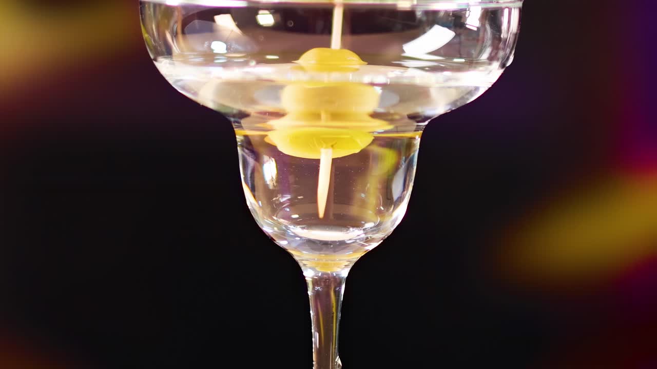 A detailed view of an olive skewer in a martini glass against a dark, colorful backdrop.