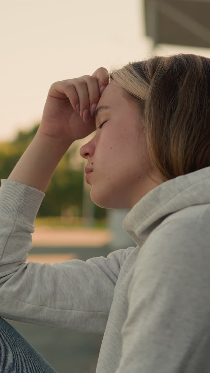 Close-up of young lady with hand on head, eyes closed, in thoughtful expression, background is softly blurred, showcasing distant building, trees, and pole
