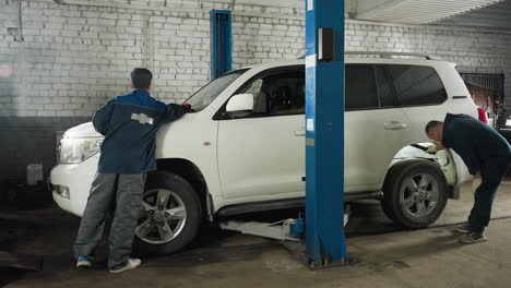 mecánicos en uniformes azules inspeccionando los neumáticos de los automóviles en su taller usando una linterna para comprobar los daños, con el vehículo levantado en un ascensor en el fondo, reparación de automóviles en progreso