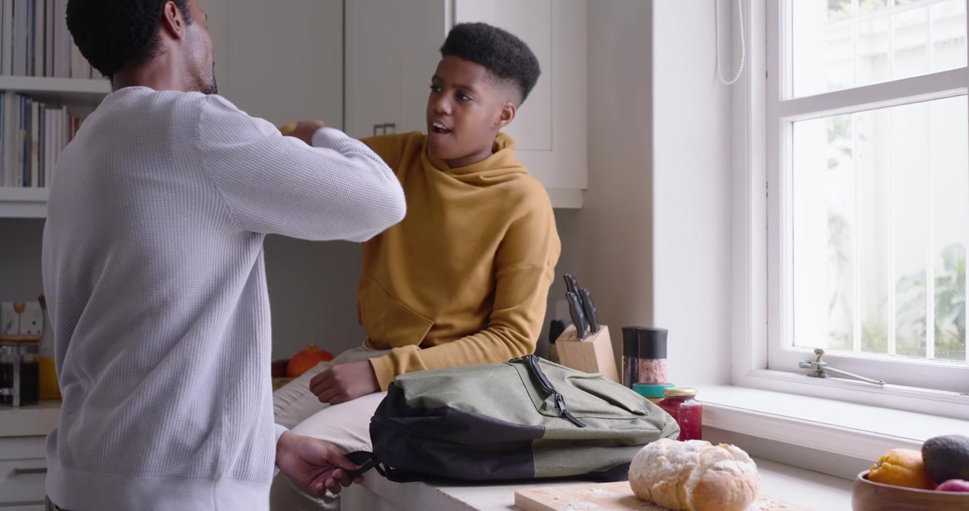 Father and teenage son packing backpack in kitchen after dad bringing lunchbox, hugging for school