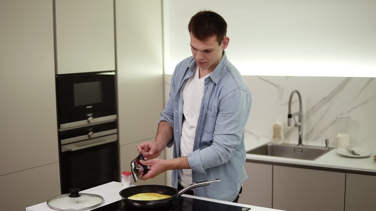 hombre en una cocina. hombre alto y guapo cocinando el desayuno, haciendo tortilla agregando especias - sal, pimienta. hombre alegre con camisa azul disfrutando de la cocina. cámara lenta