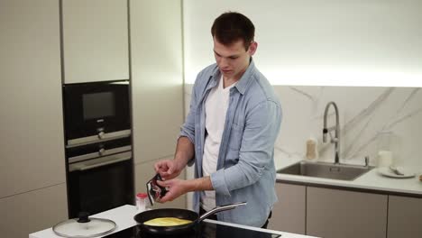 man on a kitchen. handsome tall guy cooking breakfast, making omelette adding spices - salt, pepper. cheerful man in blue shirt enjoying cooking. slow motion