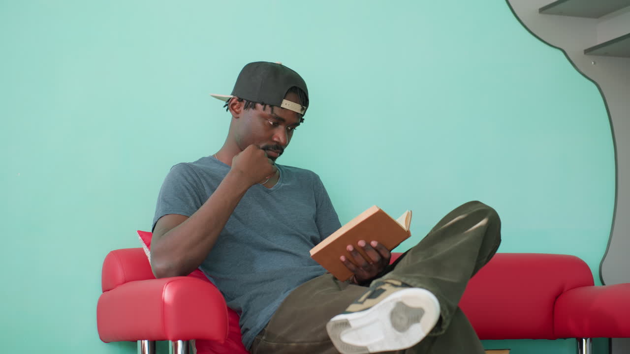 Homem de camisa casual e boné sentado em um sofá vermelho com as pernas cruzadas, segurando um livro aberto e lendo atentamente, com a mão no queixo em uma pose pensativa contra uma parede turquesa em um ambiente interno iluminado