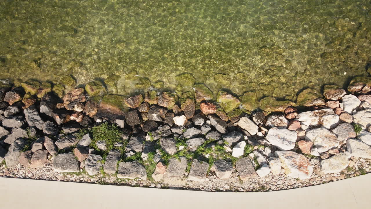 Smooth 4K drone descent to a top-down view of Lake Garda’s clear shallow water, lowering toward a rocky shoreline to reveal stones beneath the surface