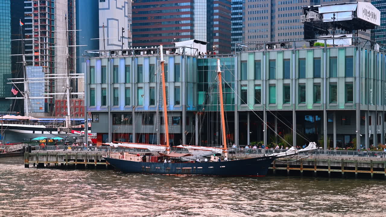 New York, USA, 1 November 2025: Historic schooner docked at a modern pier building on the Lower Manhattan waterfront. A medium perspective highlighting the contrast between the tall masts of the sailboat and the glass-clad commercial building