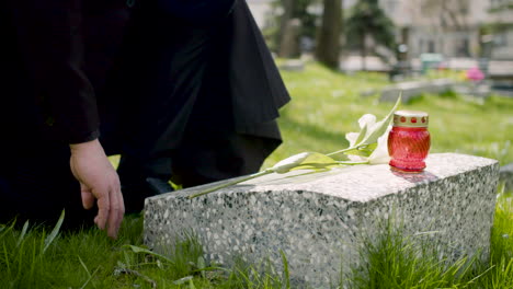 close up of an unrecognizable man in black suit kneeling and placing a white flower on a tombstone in a graveyard 1