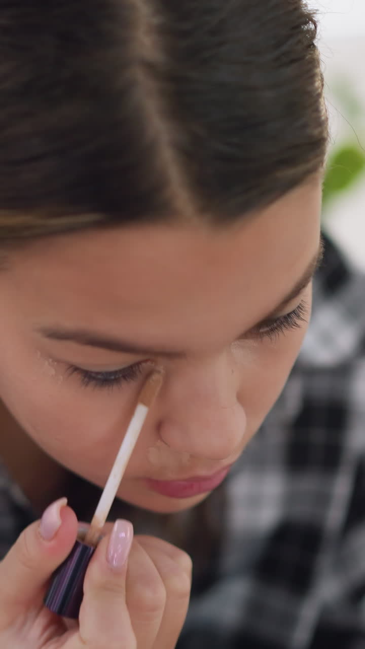 Primer plano de una adolescente terminando de aplicar el corrector de ojeras con suaves toques; primer plano de una paciente aplicando el producto sobre la piel; luz natural suave; camisa de cuadros visible; expresión tranquila y confiada; final