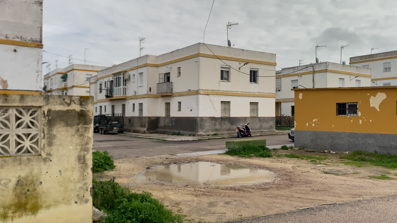 Scena di strada residenziale spagnola con semplici condomini, una grande pozzanghera su una strada non asfaltata e uno scooter parcheggiato
