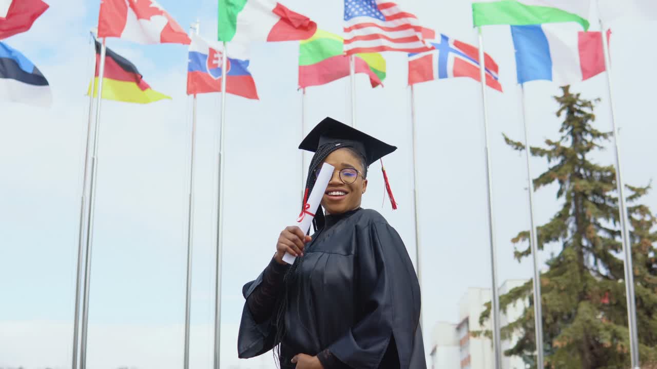 una joven afroamericana se alegra y cambia la posición de la mano en la que sostiene un diploma de educación superior. estudiante de posgrado se encuentra contra el cielo y las banderas de diferentes países