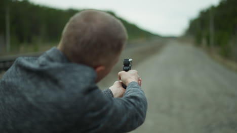 a close-up view from behind of a man in a gray jacket aiming a handgun down a deserted railway track