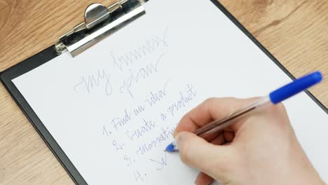 a close-up shot of a child’s hand writing the word "sell" with a blue pencil, part of a business plan, on white paper.