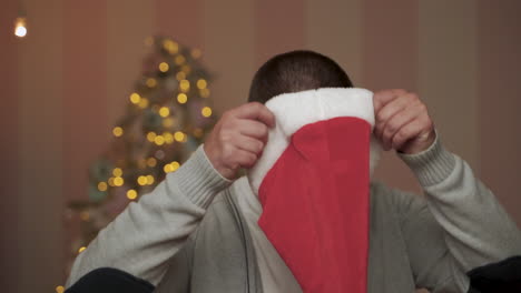 Man-Puts-On-Red-Santa's-Hat-And-Looks-At-The-Camera-With-Copy-Space