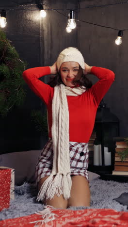 portrait of a happy model at christmas. smiling young woman putting on knitted cap and posing on camera. beautiful girl on bed on christmas decorated background. vertical video