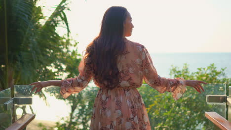 young adult asian woman from behind watching tropical ocean beach during sunset on a modern glass component balcony of resort hotel room