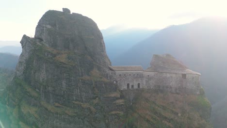 ancient fortress atop a cliff in a mountainous region, engulfed in sun rays, aerial view