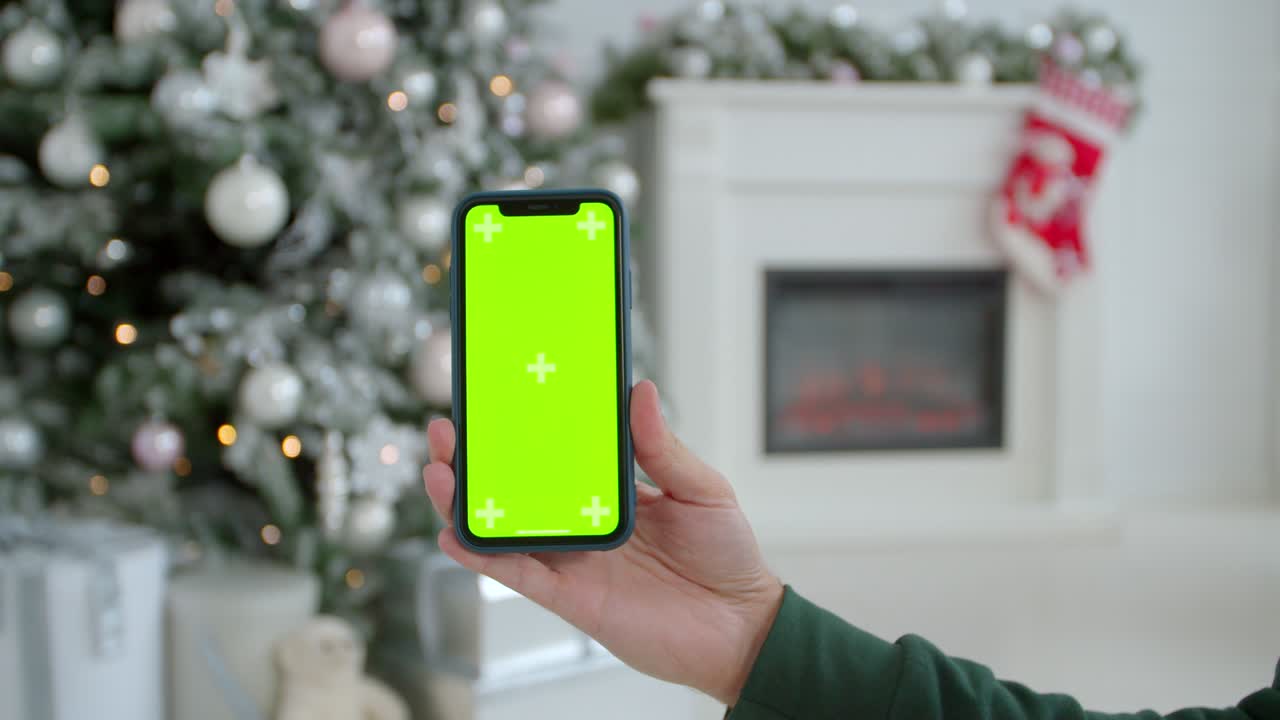 hombre con la mano sosteniendo el teléfono inteligente negro con pantalla verde en el fondo decorado de navidad. tipo de primer plano tomando fotos en el teléfono móvil permaneciendo en la sala de vacaciones festivas. tiempo de invierno.