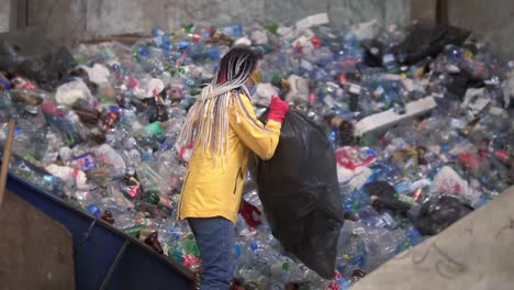 mujer-trabajadora con chaqueta amarilla y gafas de protección desempaquetando chupas negras con botellas de plástico usadas en una planta de reciclaje moderna. arrojando botellas de un saco grande en una pila en una fábrica de reciclado