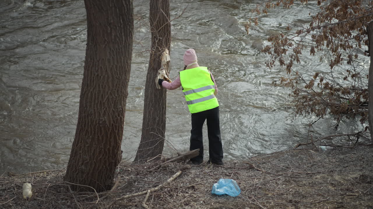 Woman in reflective safety vest stands on muddy riverbank, reaching to remove debris caught on tree trunk beside flowing water, wearing pink beanie and gloves during environmental cleanup