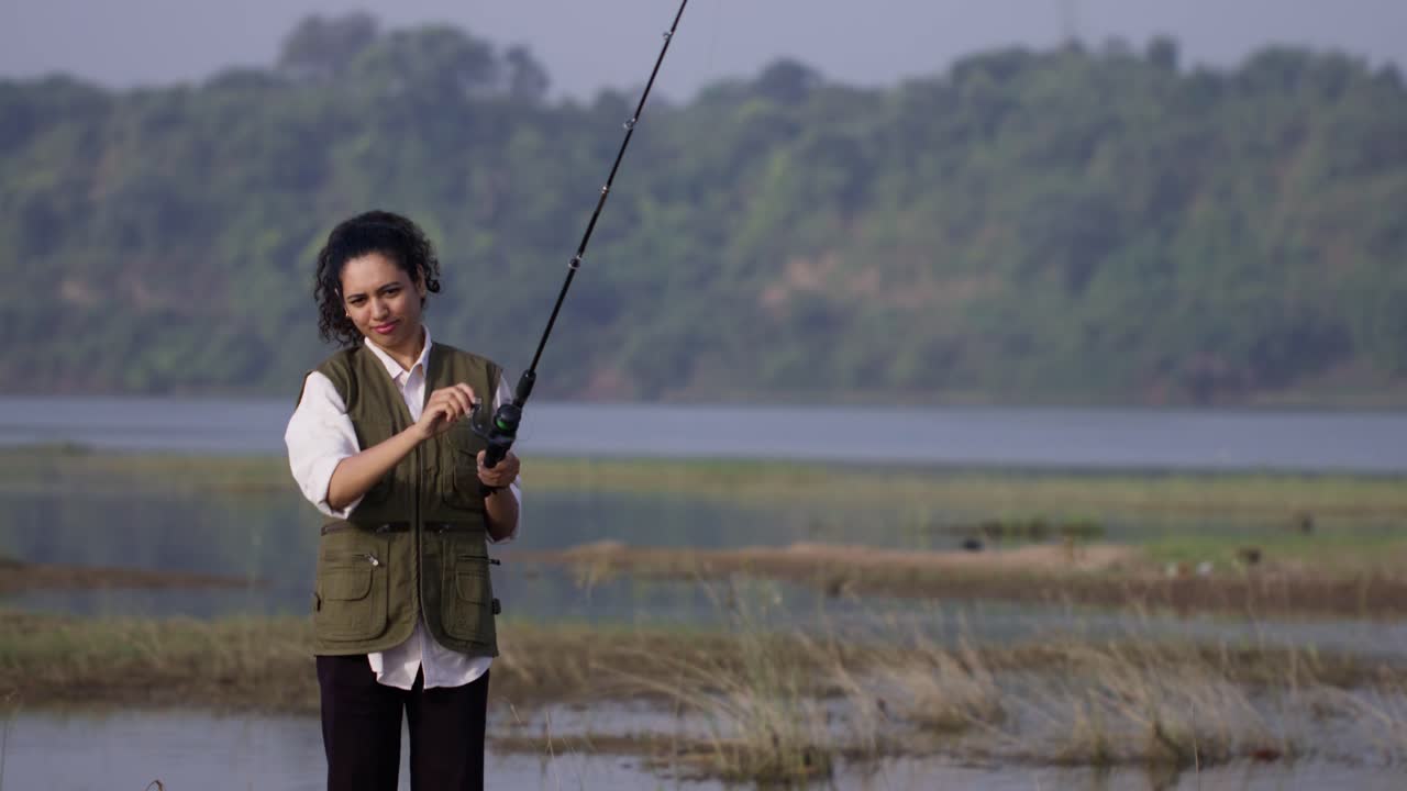 Seorang gadis Asia Selatan mengenakan rompi luar ruangan sedang memancing di tepi sungai yang tenang, melempar joran dengan fokus di bawah cahaya siang yang lembut