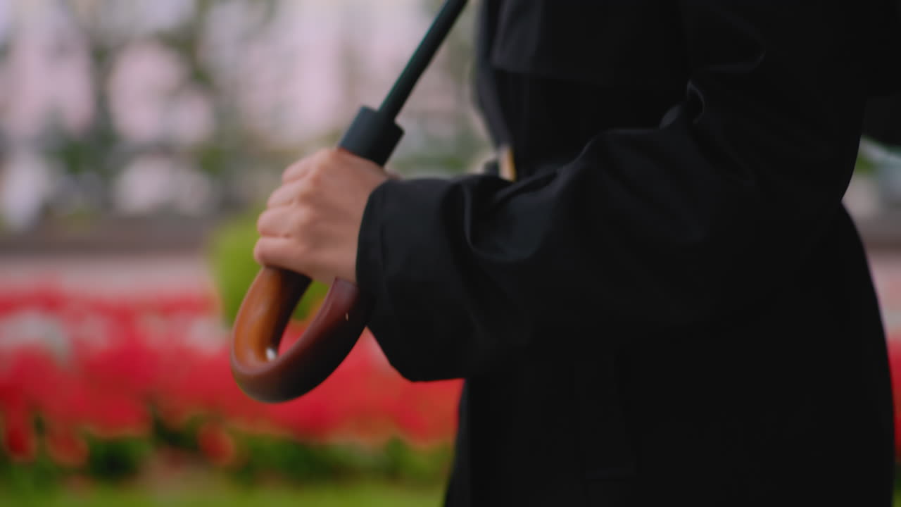 Close up of female wearing black coat holding wooden handle umbrella on rainy day outdoors with blurred background of green bushes and red flowers creating moody urban seasonal atmosphere