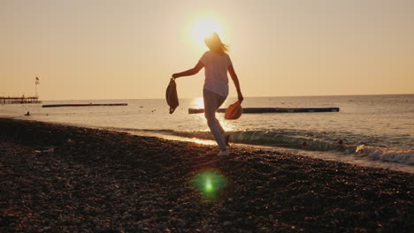 Mujer-Despreocupada-Con-Una-Mochila-Y-Un-Sombrero-En-Sus-Manos-Corre-A-Lo-Largo-De-La-Orilla-Del-Mar-Al-Amanecer-Verano-Y-Desocupa