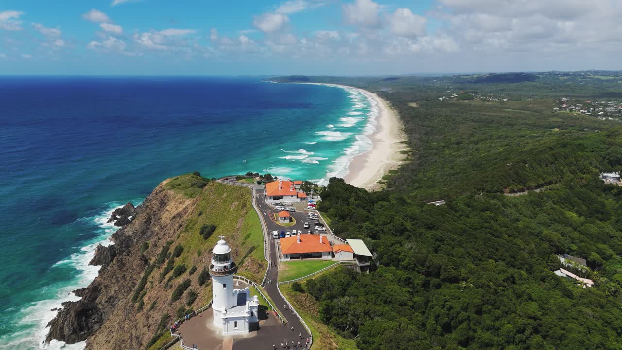 Forward drone flight hugs Cape Byron’s headland, gliding past the white lighthouse and over Byron Bay’s turquoise water, revealing New South Wales’ easternmost point at sunrise.