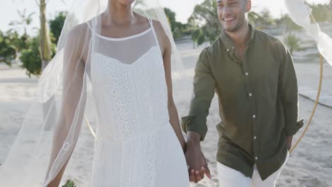 happy diverse bride and groom walking holding hands and smiling at beach wedding, in slow motion