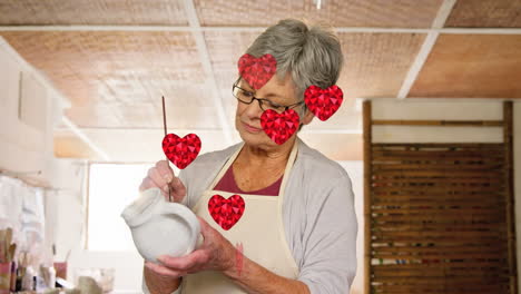 animation of heart icons over caucasian female worker painting pottery