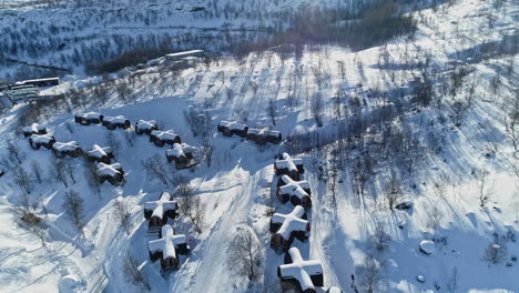 Aerial-panoramic-view-of-snow-covered-rooftops-of-cross-shape-huts-in-Snow-hotel-Kirkenes-and-sunlight-falling-on-the-landscape-symbolizing-new-day,-Norway