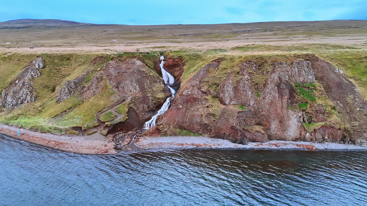 Shapeless rock sticks out from the waterscape. Drone approaches the rocky shore with a waterfall flowing from it.