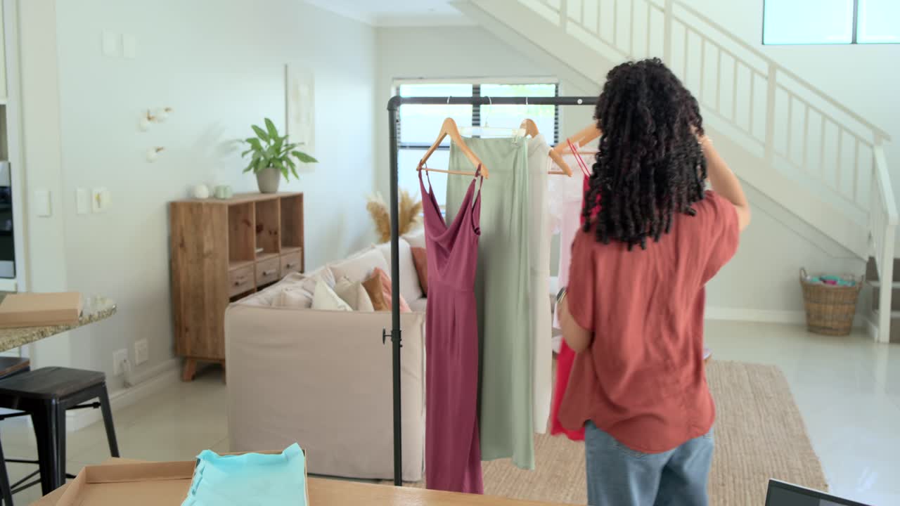 Donna che consulta riferimenti sul laptop, prende appunti e seleziona un vestito rosso per lo styling nello studio di casa.