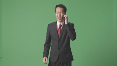 분노 한 아시아인 사업가 가 스튜디오 에서 녹색 스크린 에 서 있는 동안 휴대 전화 로 이야기 하고 있다