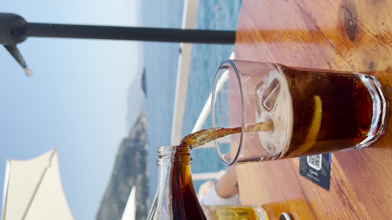 Pouring Coke with ice cubes closeup. Sea view with mountains in the distance, Croatia