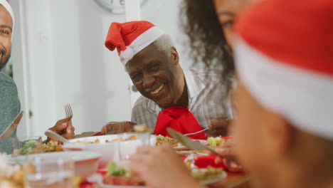 Familia-Multigeneracional-Con-Gorros-De-Papá-Noel-Disfrutando-De-Comer-Comida-Navideña-Juntos-En-Casa